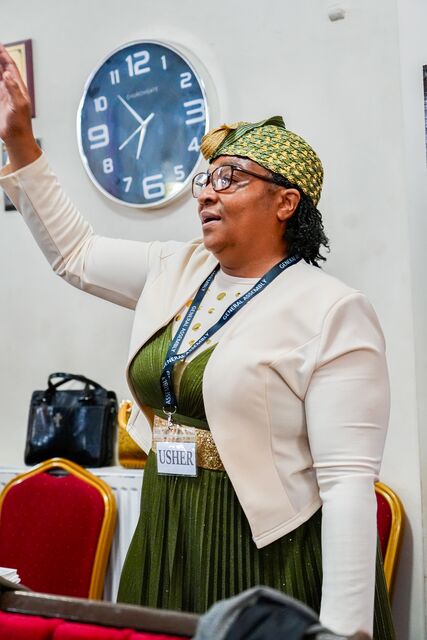 Woman in a white jacket and green dress raising one hand beside a wall clock.