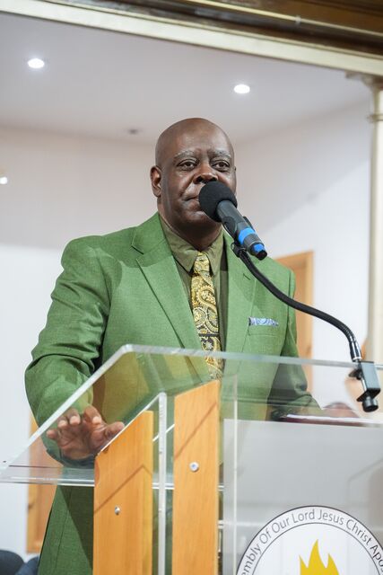 Man in a green suit speaking into a microphone behind the clear lectern.