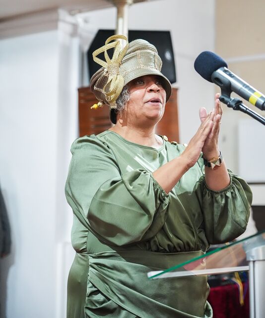 Woman in a green dress and decorative hat clapping beside a microphone.