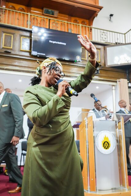 Woman in green holding a microphone and raising one hand at the lectern.