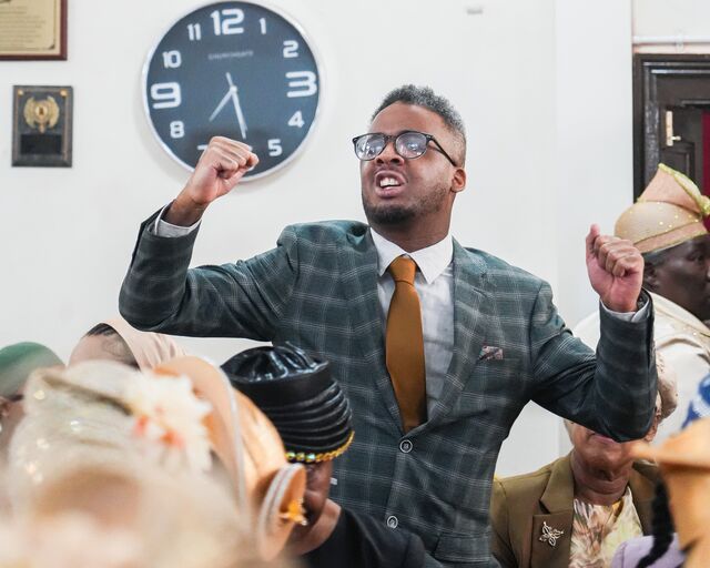 Man in a checked suit and gold tie standing with both hands raised.