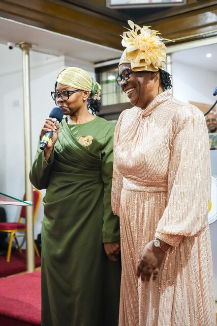 Woman in a pale pink outfit and gold hat standing beside a woman holding a microphone.