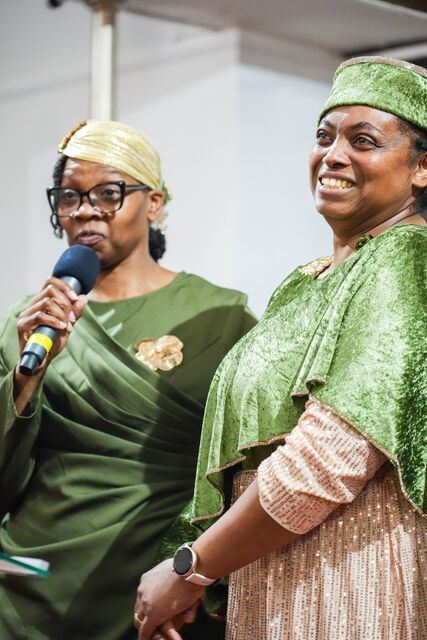 Two women in green standing together, one speaking into a microphone.
