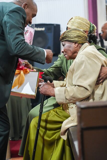 Seated woman in a cream jacket receiving an item from a man standing beside her.