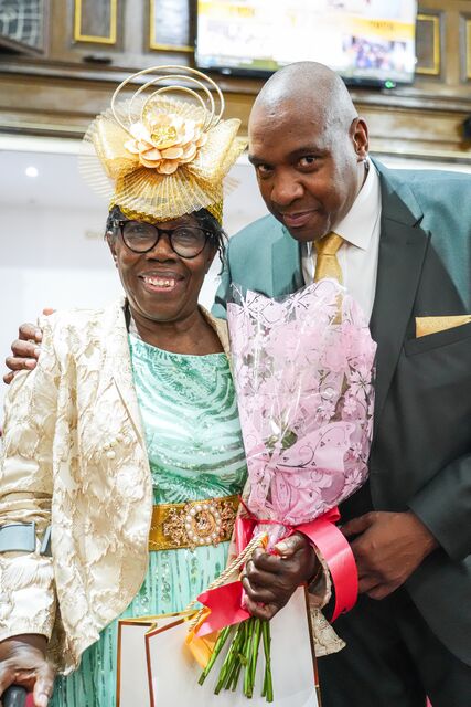 Man in a dark green suit posing beside a woman in a gold hat holding flowers.
