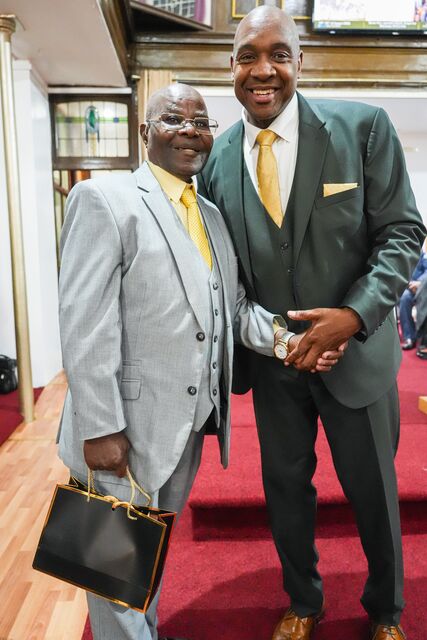 Two men posing together, one in a light grey suit and one in dark green.