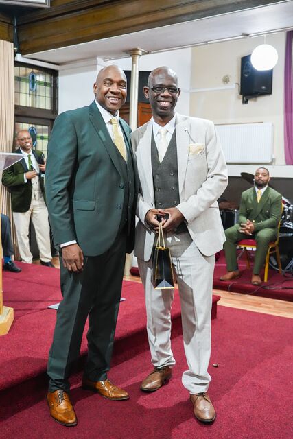 Man in a dark green suit standing beside a man in a light suit and hat.