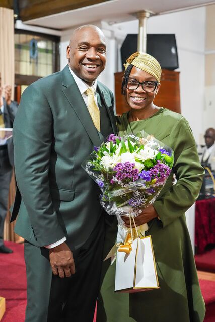 Man in a dark green suit beside a woman in green holding purple and white flowers.