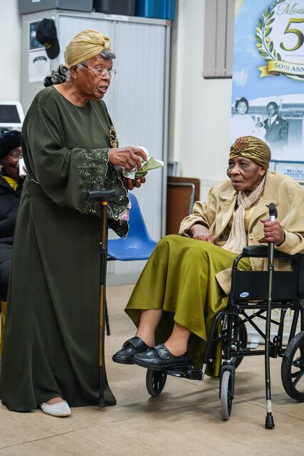 Standing woman holding a microphone beside a seated woman at the anniversary gathering.